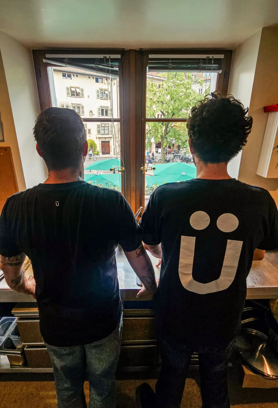 Yvan et Ben de dos, en t-shirts Ü, à une fenêtre qui donne sur la vieille ville de Genève.