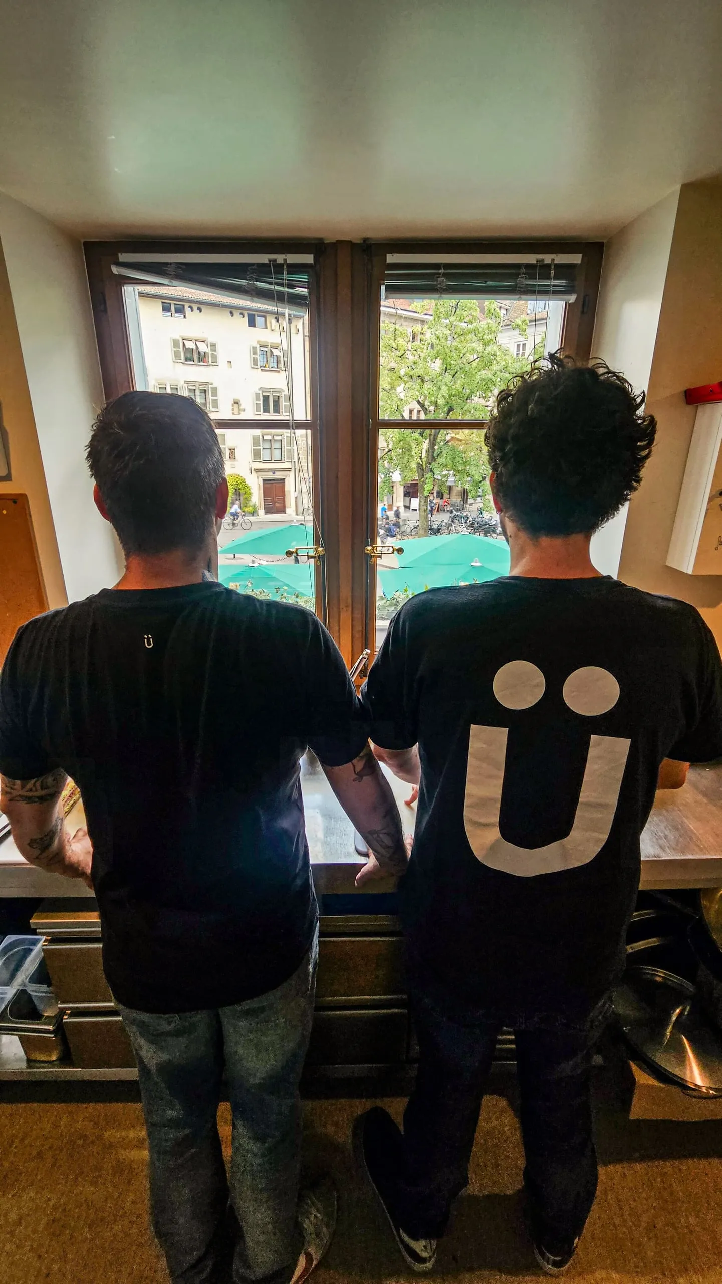 Yvan and Ben from behind, in Ü t-shirts, at a window overlooking Geneva’s old town.